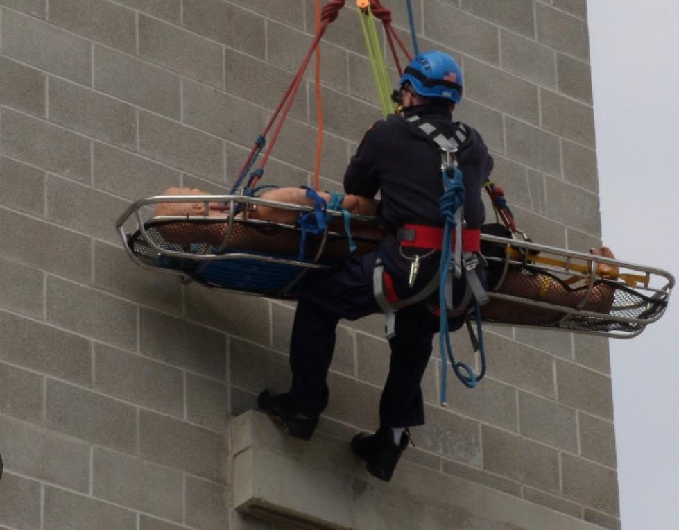 A rescuer lowering a patient in a basket stretcher down a wall